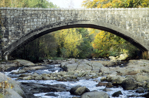 Stone bridge over the river Strontian in Scottish Highlands. Autumnal forest trees in background. Sunshine on leaves. Boulders on riverbed. Shallow waters flowing round rocks. photo