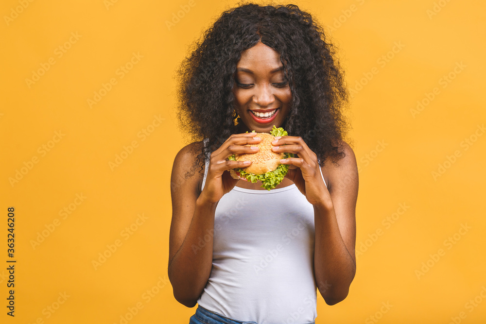 African American black beautiful young woman eating hamburger isolated on yellow background. African American black beautiful young woman eating hamburger isolated on yellow background.
