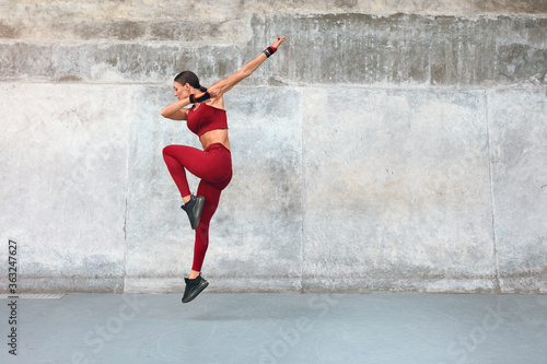 Fitness. Jumping Woman Doing Workout At Outdoor Stadium. Side View Of Fit Girl With Strong Muscular Body In Fashion Sporty Outfit Doing Exercise Against Concrete Wall. Dynamic Movement.