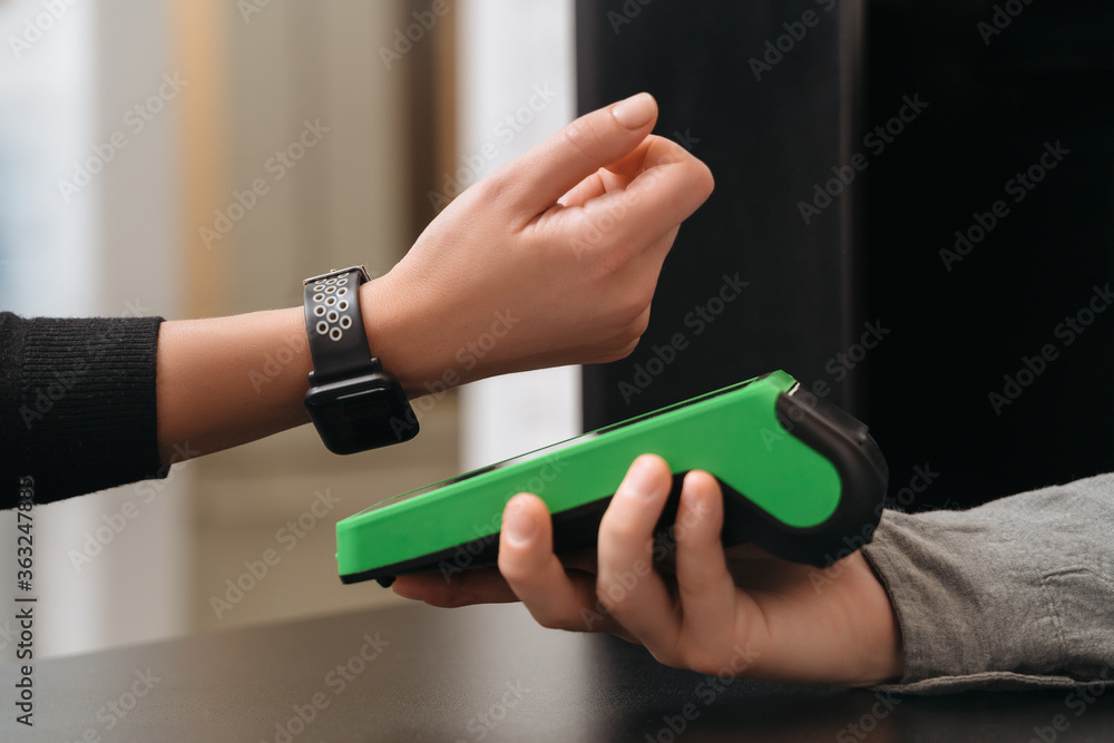 Closeup of modern lady paying via smartwatch while making purchase in shop.  Woman customer using hand watch for contactless payment via payment  terminal on counter in store. NFC technology concept Stock Photo