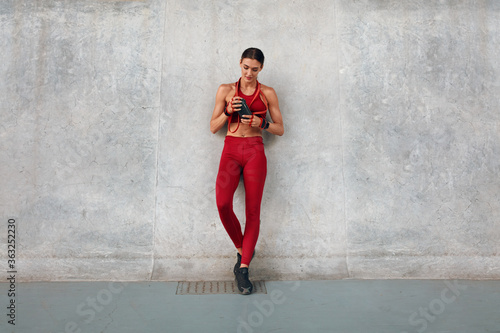 Gym. Fitness Girl With Bottle Of Protein Shake And Resistance Band Standing Near Wall. Sporty Woman In Fashion Sportswear Between Intense Cardio Workout. Outdoor Exercising As Urban Lifestyle.