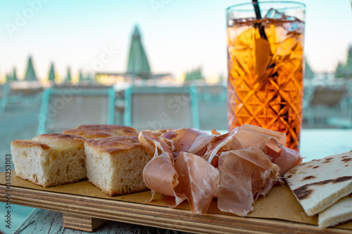 Close up of a tall glass of italian Spritz, famous cocktail with sparkling wine an orange flavored liqueur, next to a wooden tray with italian snacks like Prosciutto and Focaccia bread. photo