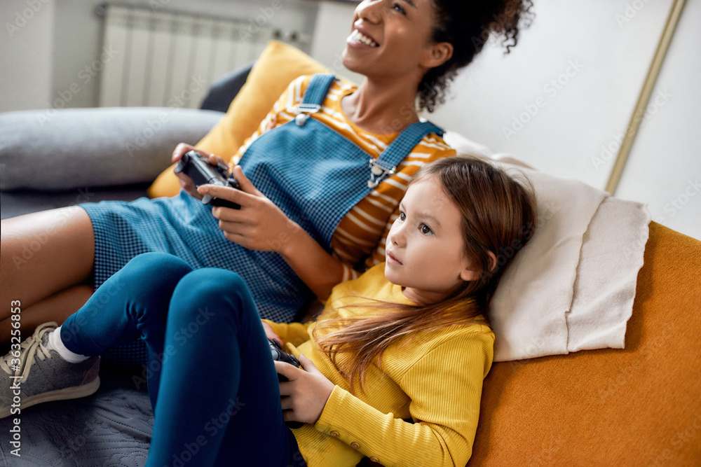 African american woman, baby sitter and caucasian cute little girl having  fun together, playing video games, sitting on the couch Stock Photo | Adobe  Stock