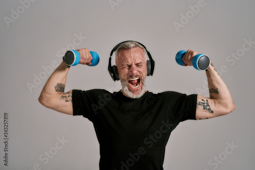 Middle aged muscular man in black t shirt and headphones closing eyes, holding blue dumbbells, posing in studio over grey background photo