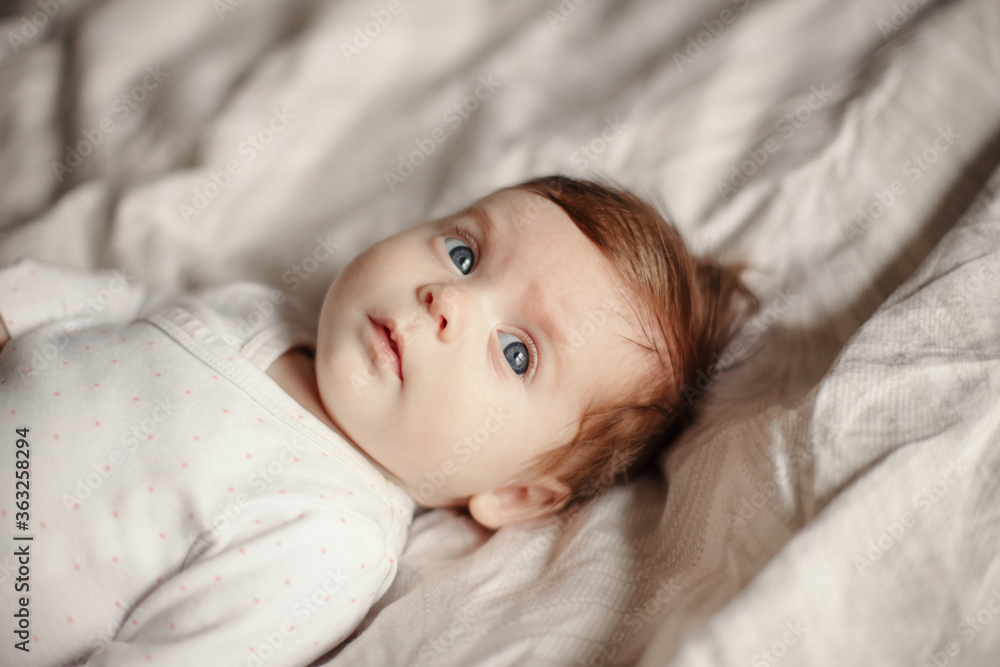 Closeup portrait of cute Caucasian newborn baby. Adorable funny child infant with blue grey eyes and red hair lying on bed looking away. Authentic childhood and lifestyle candid moments. Closeup portrait of cute Caucasian newborn baby. Adorable funny child infant with blue grey eyes and red hair lying on bed looking away. Authentic childhood and lifestyle candid moments.