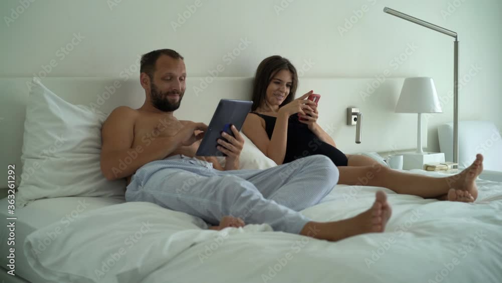 Young happy couple using smartphone and tablet computer lying on bed Young happy couple using smartphone and tablet computer lying on bed