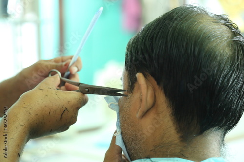 An Indian male hair dresser cutting hair in his beauty saloon with proper safety measures for Covid-19.Cutting hair of a man in mask. Hair care during Covid-19.Beauty saloon opens after lock down.