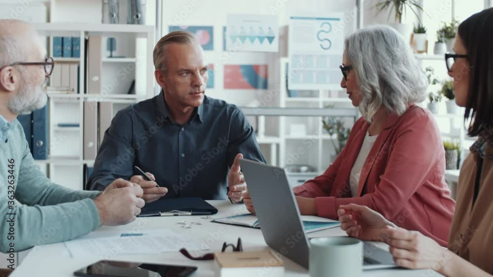Middle-aged business leader discussing project with team of mixed-aged male and female associates while holding office meeting Middle-aged business leader discussing project with team of mixed-aged male and female associates while holding office meeting