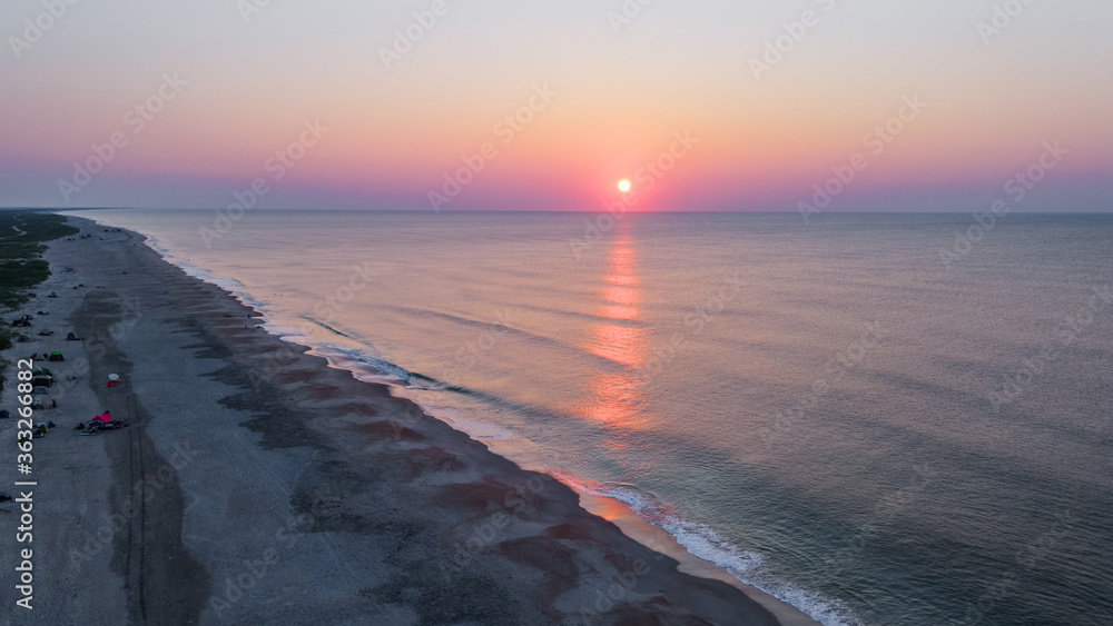 Aerial View of Beautiful Beach Sunrise overlooking Ocean Waves at Dawn with Sandy Seashore and Colorful Pastel Skyline and Reflection of Sun Rays in Water on Coastline