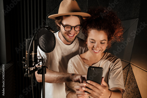 Beautiful couple of stylish singers happily using phone working together in sound recording studio photo