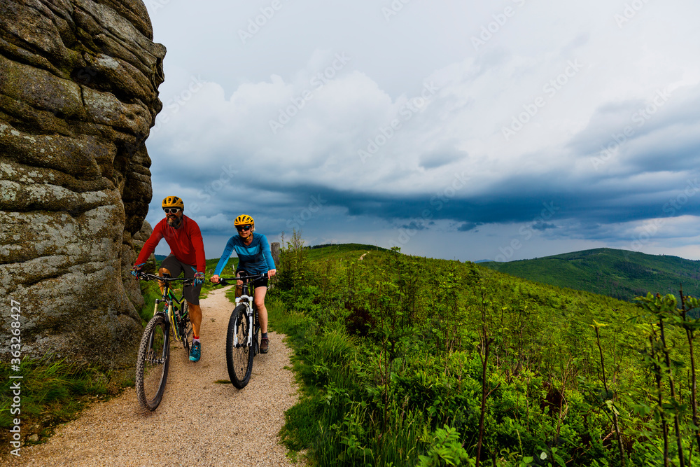 Fototapeta premium Cycling woman and men riding on bikes at sunset mountains forest landscape. Couple cycling MTB enduro flow trail track. Outdoor sport activity.