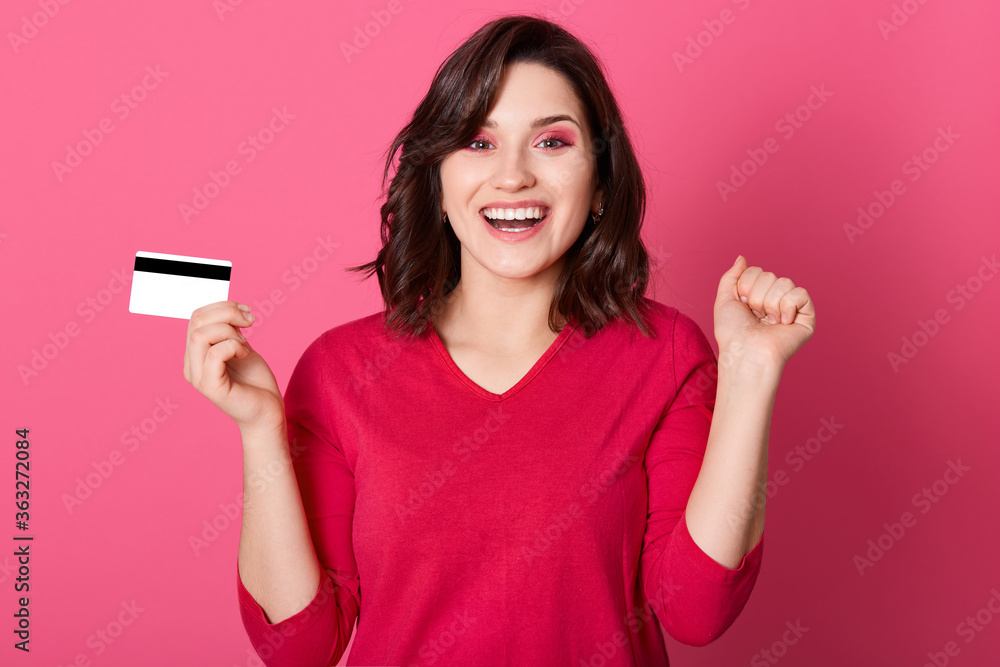 Young brunette woman shouting with happy expression and keeping fists clenched, celebrating success, holding credit card, wearing red casual shirt, standing against rosy wall. Young brunette woman shouting with happy expression and keeping fists clenched, celebrating success, holding credit card, wearing red casual shirt, standing against rosy wall.
