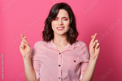 Close up photo of beautiful amazing lady keeping fingers crossed, stands with closed eyes, asking for win, female with short hairstyle, wearing casual shirt isolated rose background.