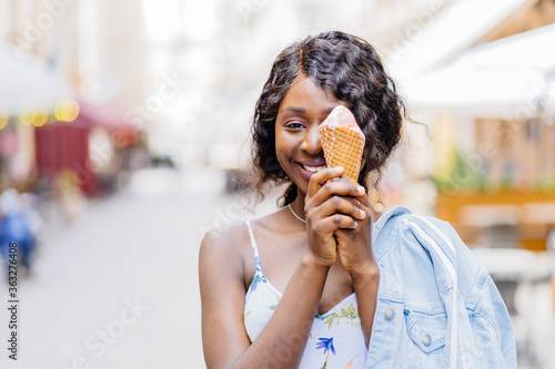 Attractive african playful woman on the street having fun and eating ice cream. Dreaming outdoor summer portrait of millennial black girl in straw hat  blue jeans jacket enjoying summer.