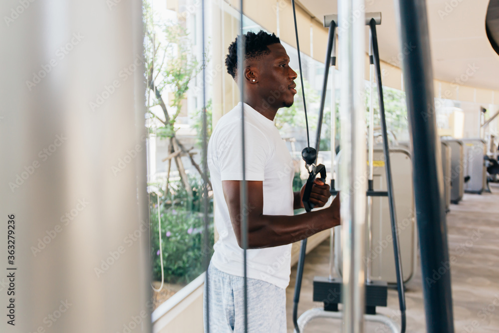 Muscular African American sportsman doing cable fly with exercise machine standing against window during training in gym. Muscular African American sportsman doing cable fly with exercise machine standing against window during training in gym.