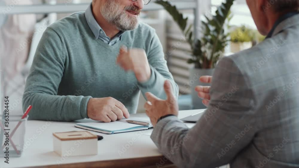 Midsection shot of senior bearded businessman smiling and speaking with male associate while sitting at office desk Midsection shot of senior bearded businessman smiling and speaking with male associate while sitting at office desk
