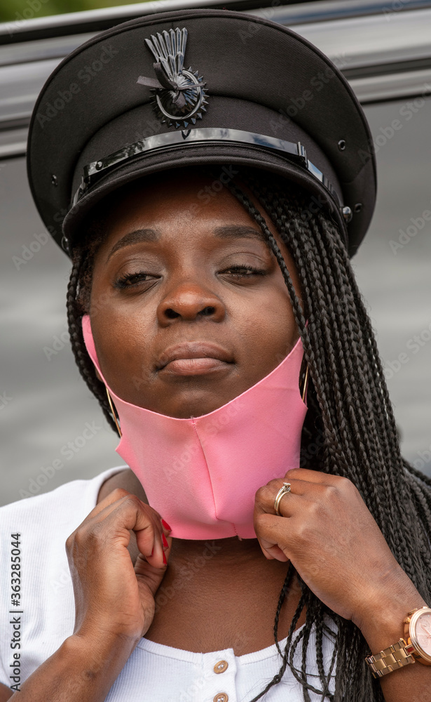 Fototapeta premium England, UK. 2020. Portrait of a woman chauffeur with uniform cap and braided hair adjusting her pink coloured facemask during the Covid-19 outbreak