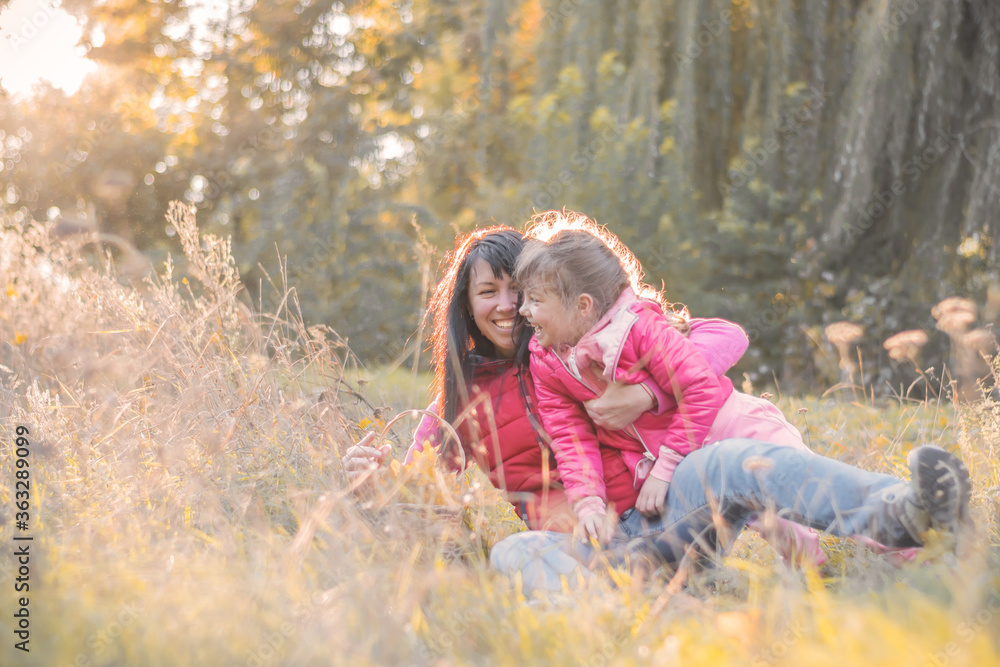 Mom and daughter fall to the ground in the autumn park. The family plays having fun and lying around in the grass. Cheerful girls actively relax in nature. Mom and daughter fall to the ground in the autumn park. The family plays having fun and lying around in the grass. Cheerful girls actively relax in nature.