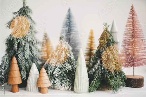 Christmas little trees and golden lights bokeh on white background. Winter magic forest, festive modern decor. Happy holidays. Miniature ceramic, wooden, snowy and handmade pine trees.
