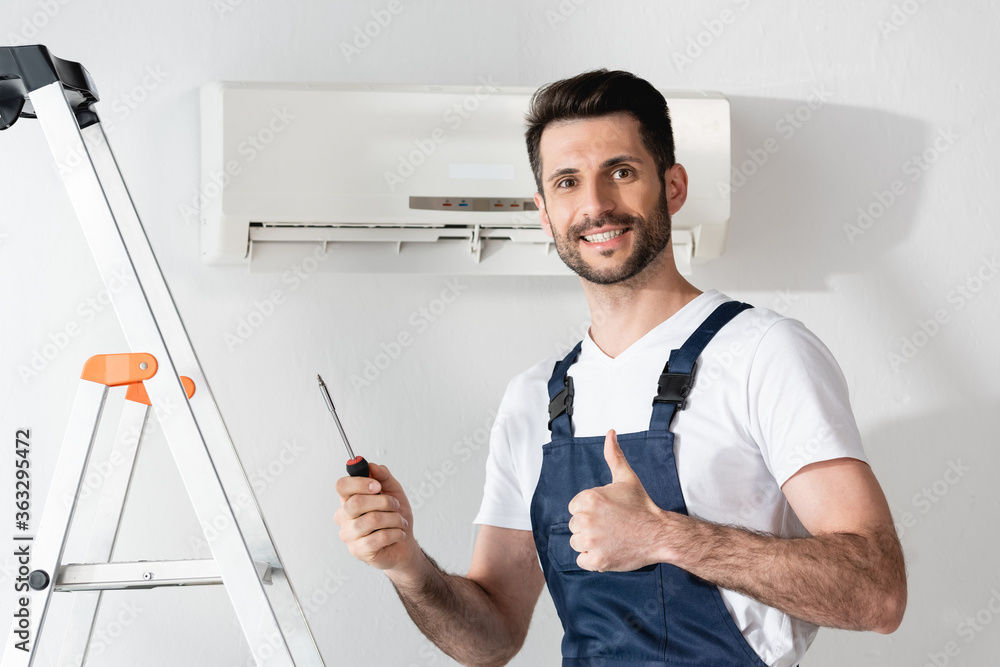 happy repairman showing thumb up and holding screwdriver while standing near stepladder and air conditioner