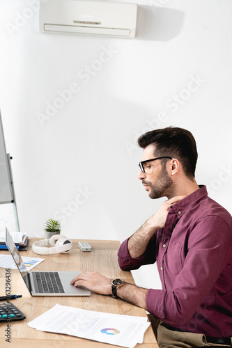 young businessman working on laptop and touching neck while suffering from heat under air conditioner