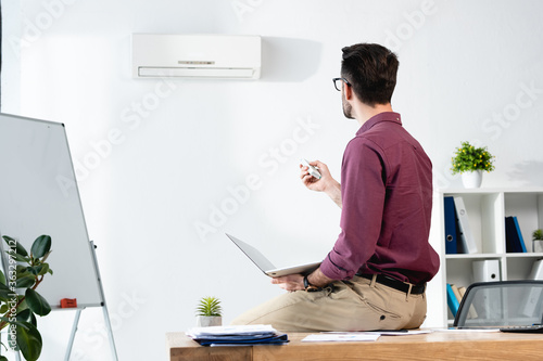 businessman sitting on desk with laptop and switching on air conditioner with remote controller