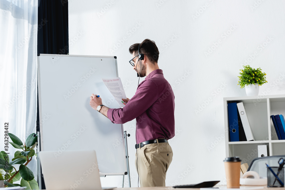 young businessman in headset writing on flipchart while holding document near desk with laptop