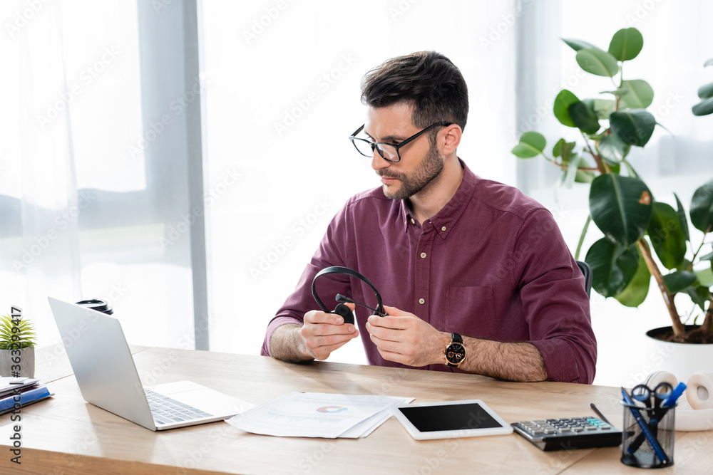young, serious businessman holding headset while looking at laptop during video call young, serious businessman holding headset while looking at laptop during video call