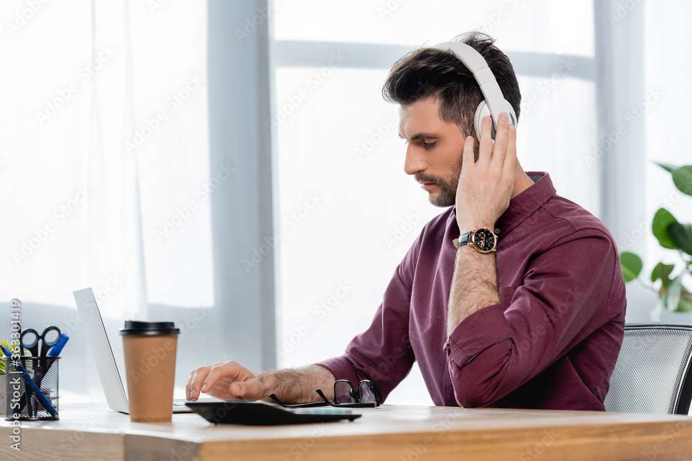 concentrated businessman in wireless headphones typing on laptop near coffee to go concentrated businessman in wireless headphones typing on laptop near coffee to go