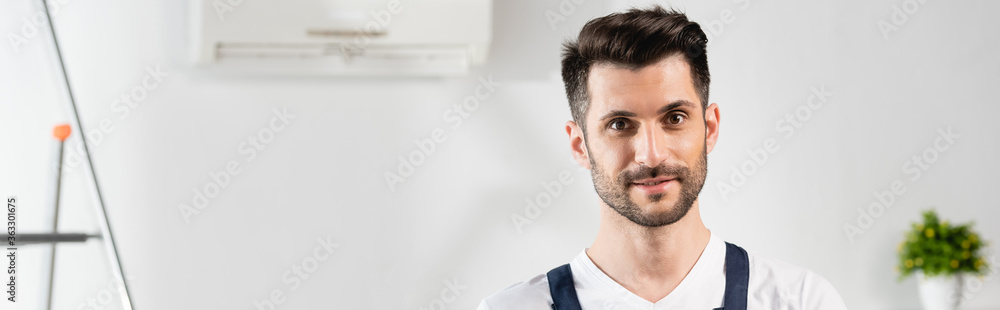 Fototapeta premium horizontal image of handsome, smiling repairman looking at camera near air conditioner on wall