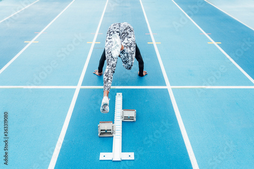 Back view of young strong sportswoman beginning to run fast from starting blocks during track and field workout on stadium