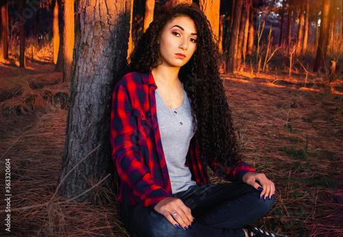 Menina adolescente negra com cabelos cacheados volumosos, sentada na floresta, ao pôr do sol, encostada em uma árvore, vestindo camisa xadrez. Beleza afro, beleza negra.
Clima de outono photo