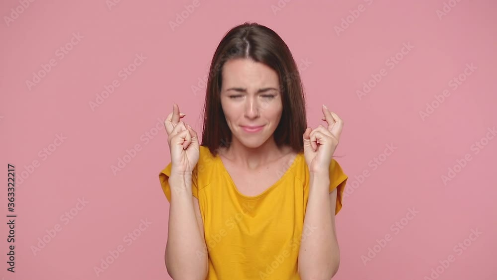 Young woman in yellow t-shirt isolated on pastel pink background studio. People lifestyle concept. Look camera bit lips hands folded in prayer beg about something good keep fingers crossed making wish