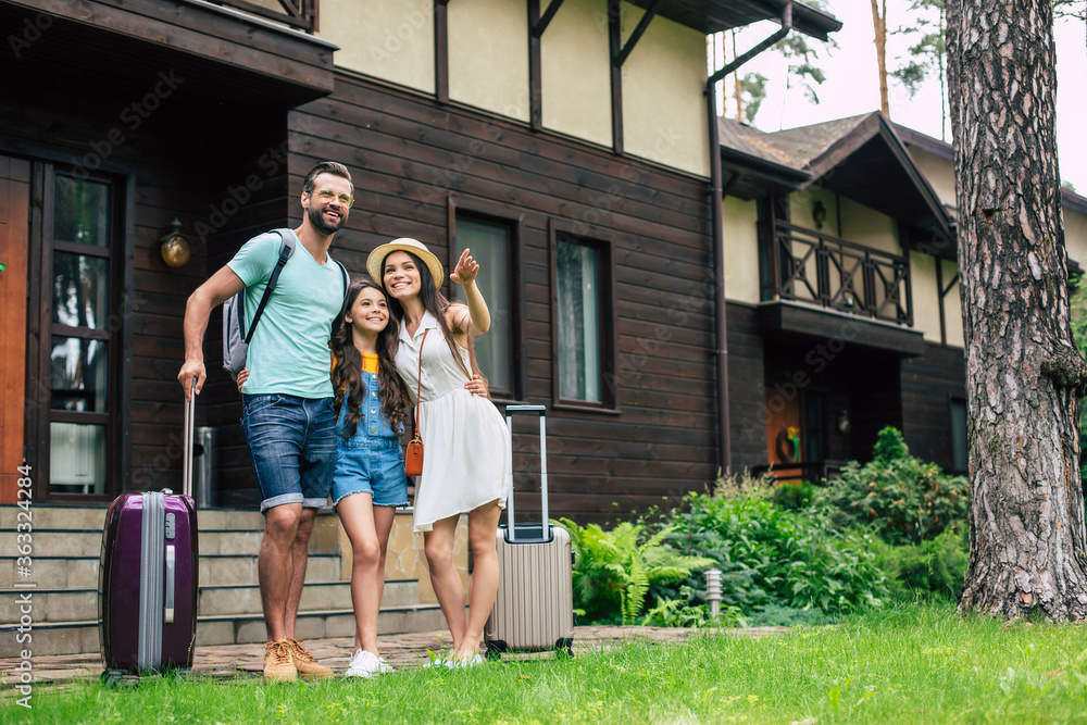 Photo of a beautiful young tourist family on vacation while them standing on forest hotel background and waiting to get settled in the room with backpacks and suitcases.