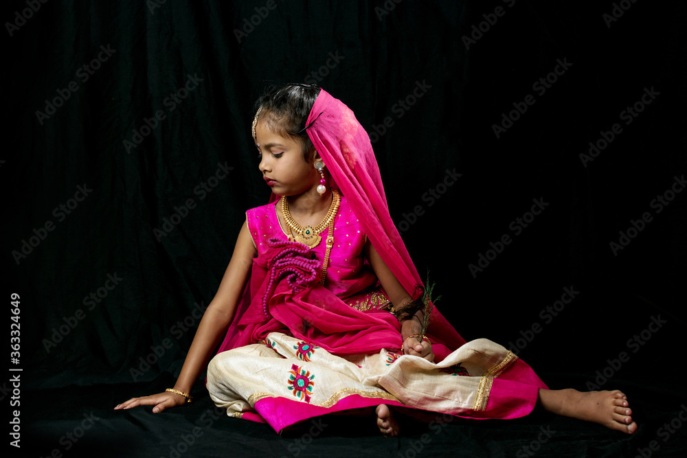 Indian girl child wearing ethnic costume on Krishna Jayanti festival sitting like Radhe isolated in black background. Indian girl child wearing ethnic costume on Krishna Jayanti festival sitting like Radhe isolated in black background.