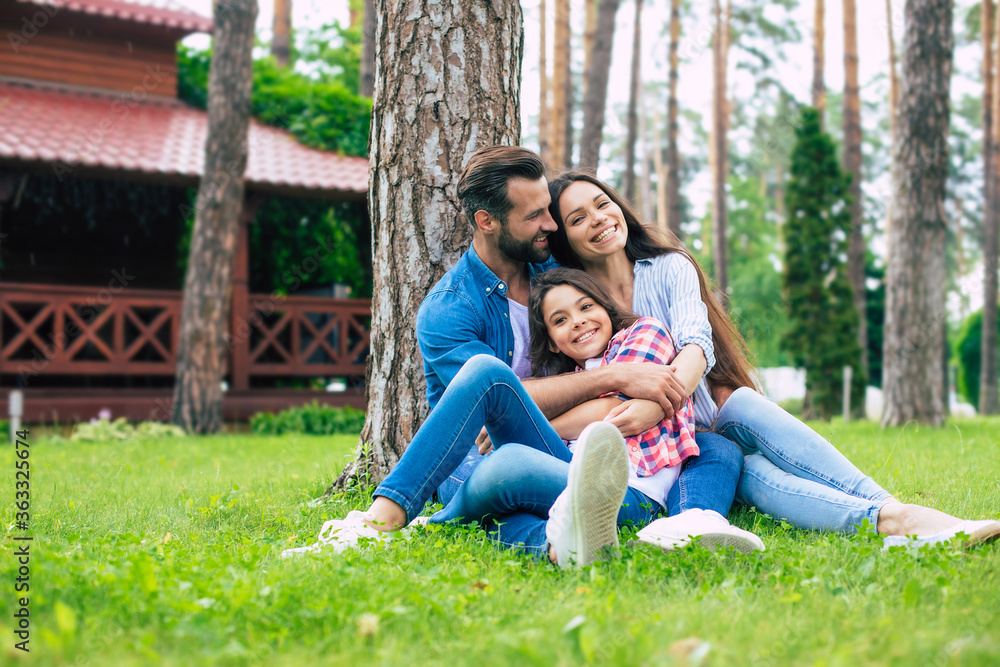 Beautiful happy family while sitting together on the grass and hugging each other, relaxing outdoors on suburban house background. Beautiful happy family while sitting together on the grass and hugging each other, relaxing outdoors on suburban house background.