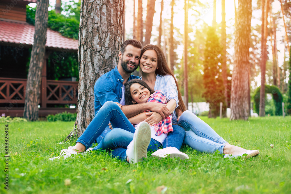 Beautiful happy family while sitting together on the grass and hugging each other, relaxing outdoors on suburban house background.