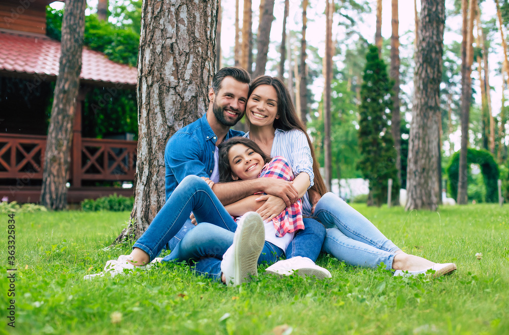 Beautiful happy family while sitting together on the grass and hugging each other, relaxing outdoors on suburban house background.