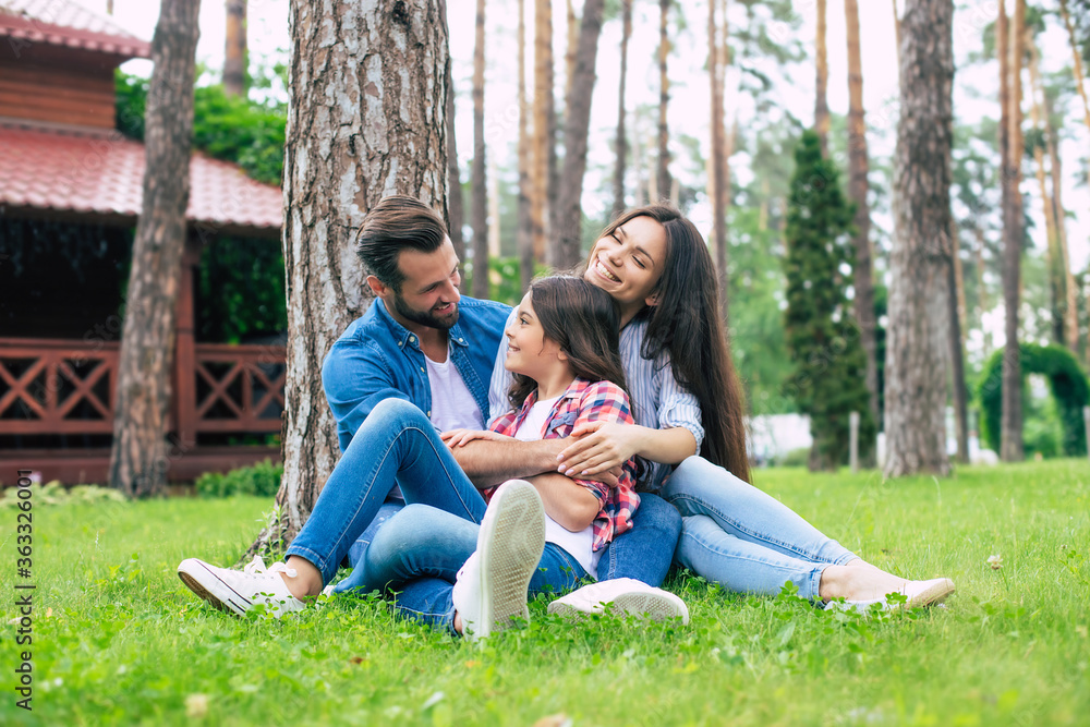 Beautiful happy family while sitting together on the grass and hugging each other, relaxing outdoors on suburban house background. Beautiful happy family while sitting together on the grass and hugging each other, relaxing outdoors on suburban house background.