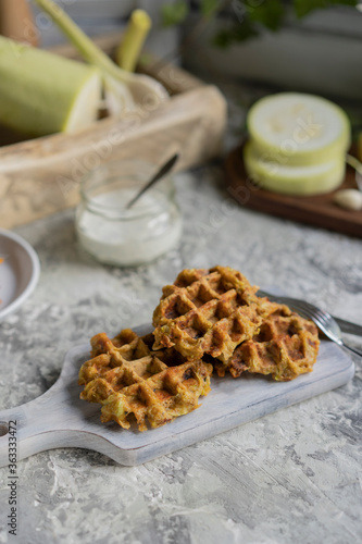 Savory squash or zucchi waffles with cheese, herbs and white sauce. Homemade baked waffles on grey textured table at kitchen, natural light, organic products on background. Selective focus, copy space photo