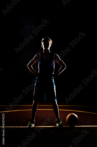 Basketball. The silhouette of a teenage Boy in blue sportswear confidently poses with his hands on his hips. The ball is on the ground. Black background. Concept of sports games