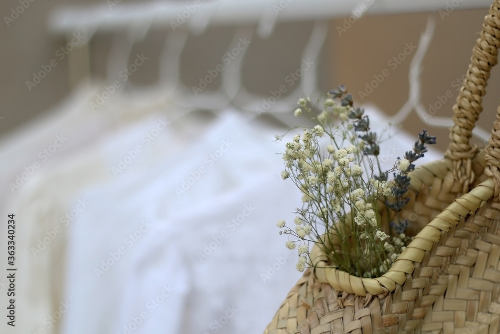 Clothing rack with white and neutral garments and wicker bag with flowers. Selective focus, gray concrete background.