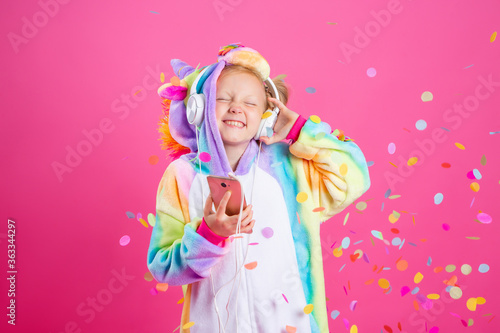 Happy little blonde girl in unicorn kigurumi listens to music holding in hand smatrphone on a pink background