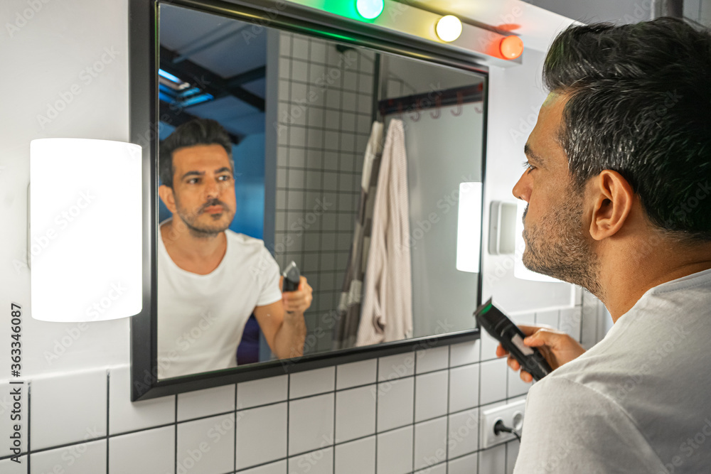Fototapeta premium handsome bearded man is shaving his face with trimmer machine in front of bathroom mirror