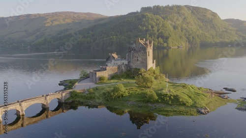 Aerial drone shot tracking around the stunning Eilean Donan Castle in Scotland, with the lake surrounding it and mountains in the background, and the reflection in the water.