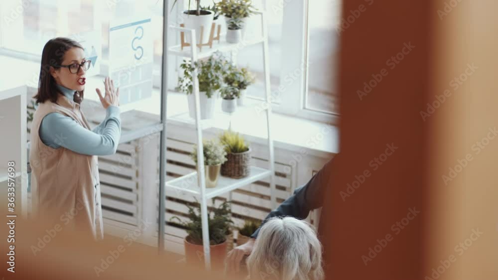 High angle shot of young female coach pointing at posters on glass wall and explaining presentation to business team during corporate education seminar in office High angle shot of young female coach pointing at posters on glass wall and explaining presentation to business team during corporate education seminar in office