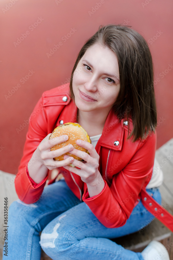 Young girl eating a burger outdoors. Girl with pleasure eats fast food on the street. Attractive girl bites a burger