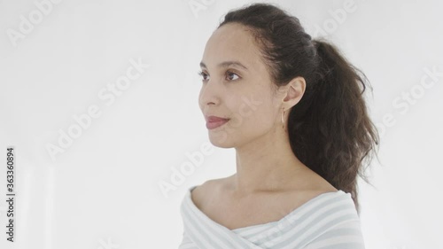 Beautiful young dark-haired female in white stripped longsleeve smiles contentedly and looks straight away. 360 degree tracking arc shot in 4K.