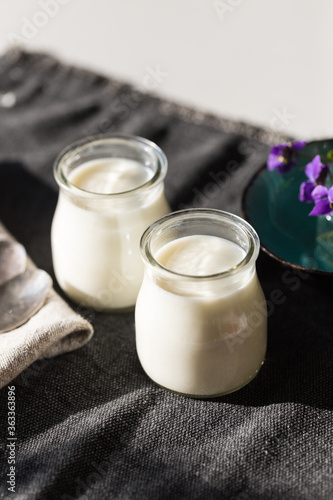 Two glass jars with natural homemade yogurt, spoons, little vase with violets on black fabric on white wooden table.