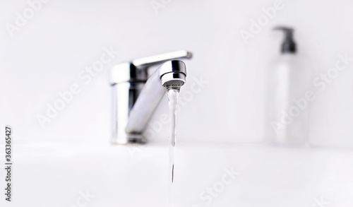 Faucet, water flow, and a blurred bottle of sanitizer on the background. Antiseptic dispensers near water tap sink with faucet, washing hands with soap to protect from coronavirus.